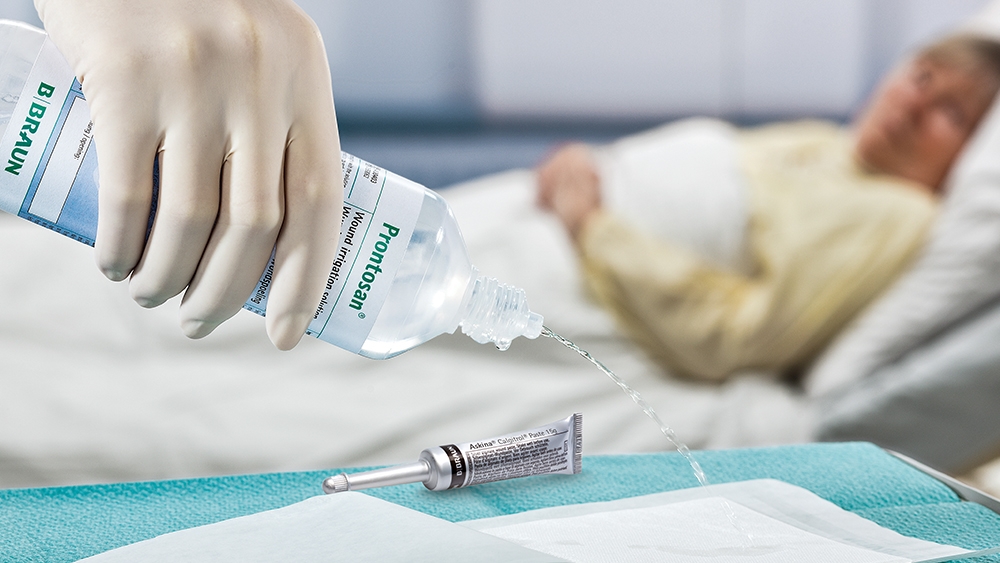 Em primeiro plano, uma mão usando uma luva médica derrama a Solução de Irrigação Prontosan em uma almofada sobre a mesa ao lado de um tubo Askina Calgitrol. Ao fundo, uma paciente está deitada em uma cama de hospital.