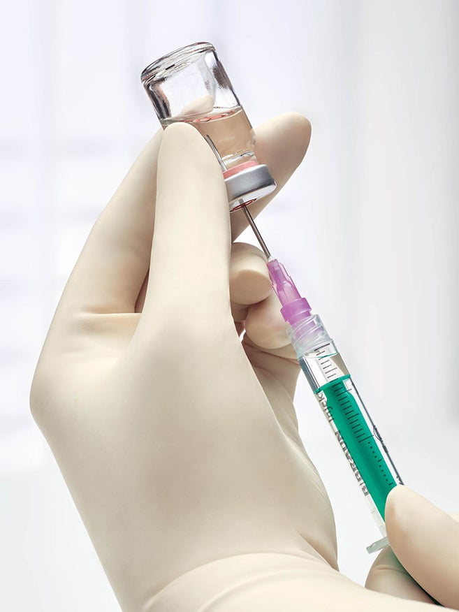 Close de uma mão com luva retirando solução de um frasco transparente com uma seringa, ilustrando a preparação estéril de medicamentos com seringa e frasco. 