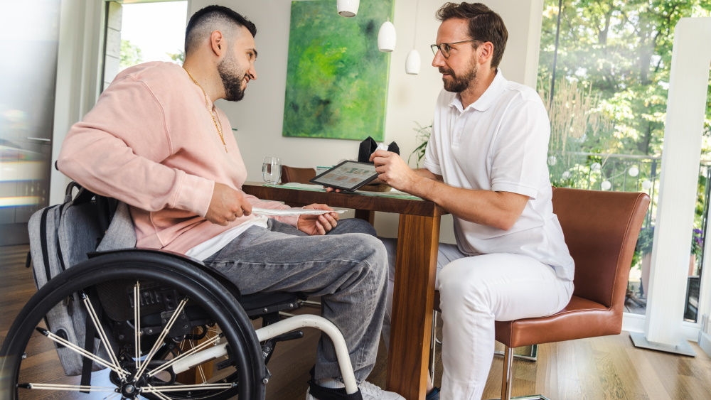 Um homem está sentado em uma cadeira de rodas, segurando um cateter nas mãos. À sua frente, um especialista em saúde hospitalar está dando instruções usando um tablet