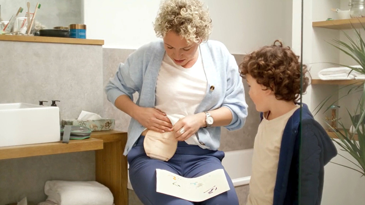 Uma mulher de camisa branca sentada no banheiro com uma bolsa de ostomia no estômago e um manual nos joelhos. Uma criança está ao lado dela e olha para sua bolsa de ostomia. 