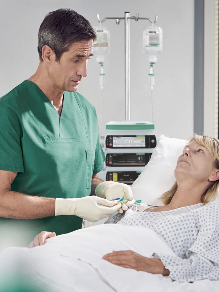 Uma mulher loira está deitada em uma cama de hospital. Ela está conectada a uma infusão, e uma bomba Spaceplus pode ser vista ao fundo. Um médico de cabelos escuros, vestindo uma bata cirúrgica verde, está ao lado do leito. Ele está regulando a administração do medicamento para dor por infusão e conversando com a mulher.
