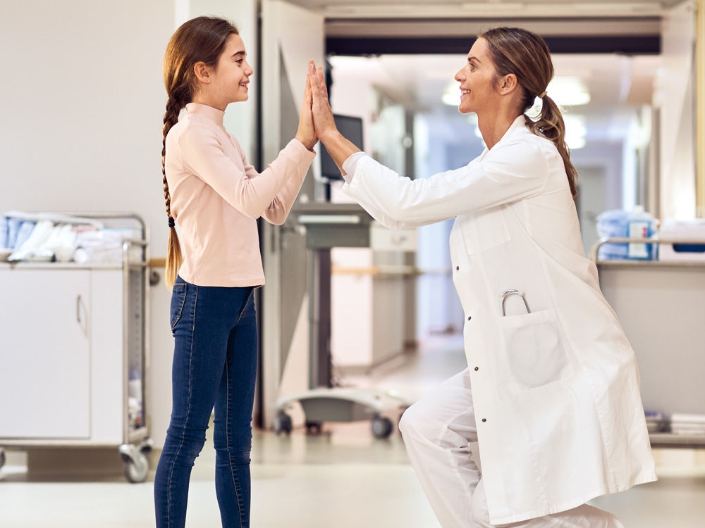 Uma menina de cabelos escuros e um médico de cabelos escuros estão de pé, um em frente ao outro, no corredor de um hospital, ambos sorrindo. O médico se agachou para ficar na altura dos olhos do pequeno paciente. Eles se cumprimentam batendo as duas mãos.