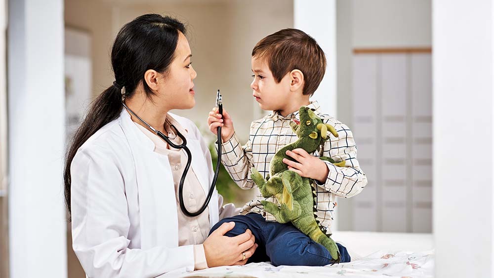 Uma profissional de saúde está conversando com um garotinho que está segurando um dinossauro de brinquedo. O menino parece empenhado e curioso, enquanto a profissional de saúde tem uma expressão calorosa e amigável