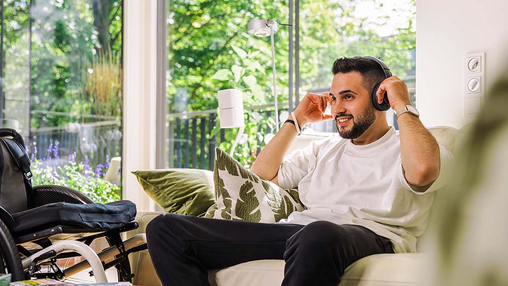 À direita, um homem está sentado no sofá com fones de ouvido, segurando as mãos sobre os fones. À sua esquerda há uma cadeira de rodas