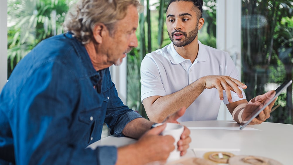 Um homem mais velho com café e um jovem com um tablet sentam-se em uma mesa e conversam
