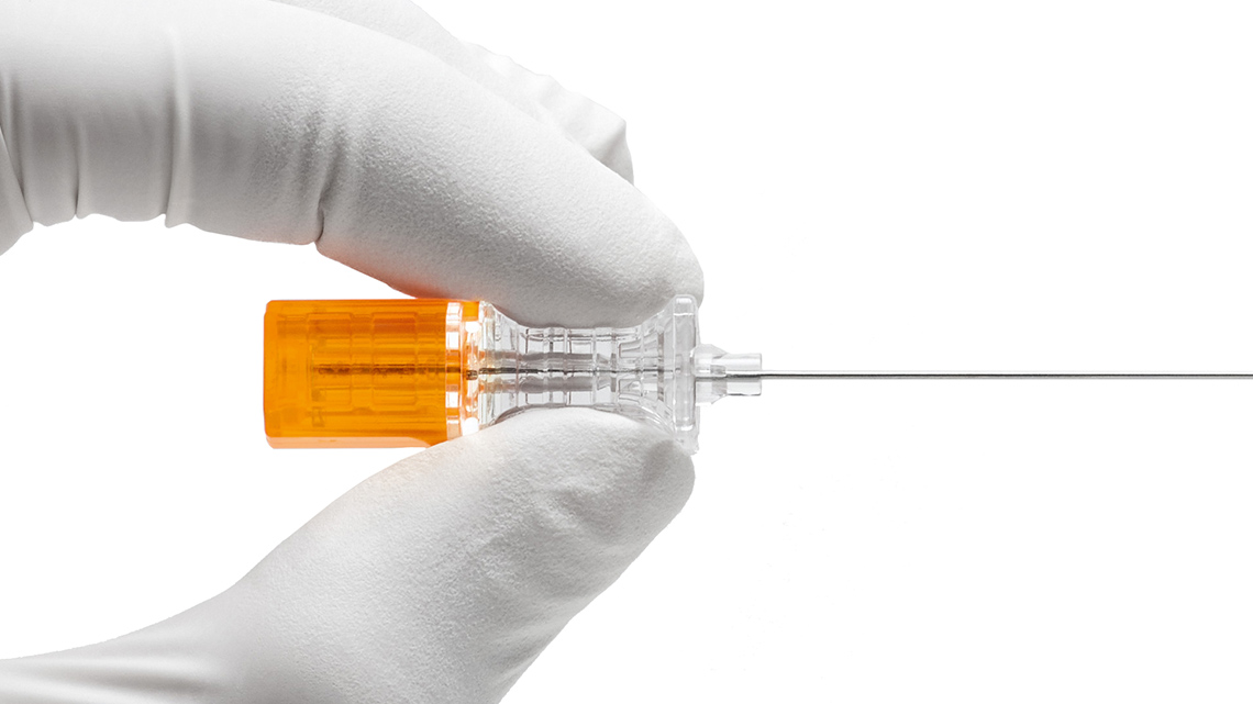 Close-up de uma mão enluvada segurando uma agulha espinhal Pencan®, com canhão laranja e corpo transparente, utilizada em procedimentos médicos