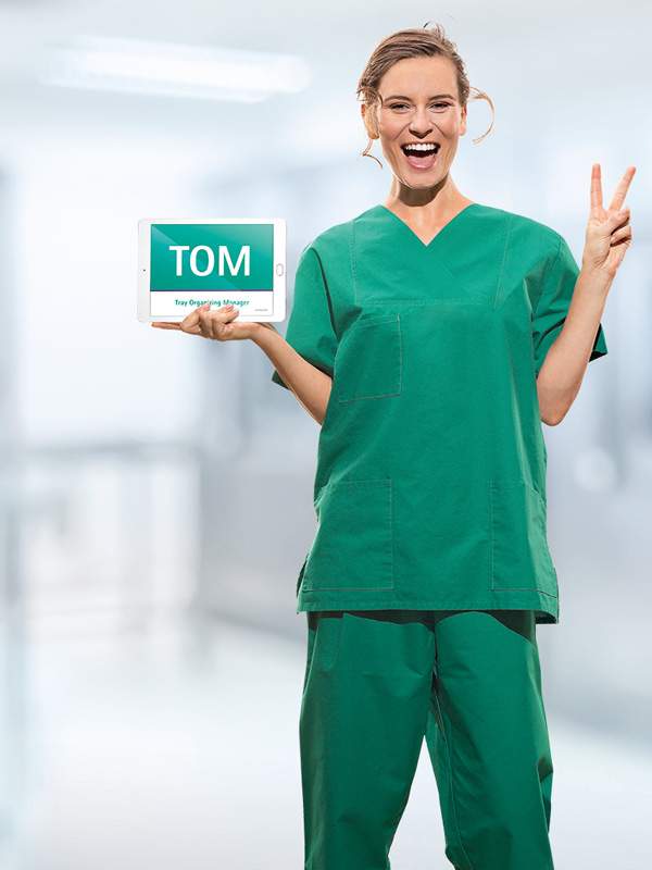 A female medical worker, dressed in medical attire, is holding a tablet with the abbreviation ‘TOM’ displayed on it. She is also showing a ‘V’ sign with her fingers