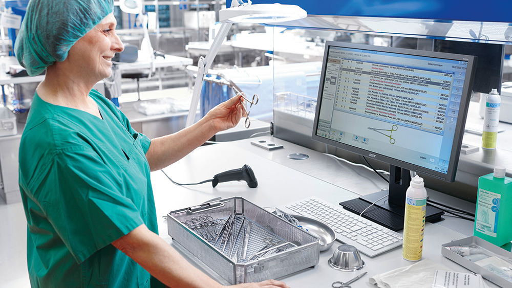 Uma profissional de saúde vestida com uniforme cirúrgico está ao lado de um computador, segurando um instrumento cirúrgico e comparando-o com uma imagem na tela