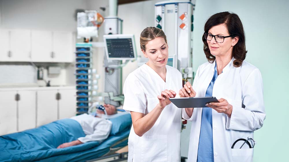 Um médico e uma enfermeira estão discutindo a dosagem da infusão inteligente da B. Braun, que está sendo inserida diretamente de um tablet. O paciente na unidade de UTI pode ser visto em segundo plano, conectado a uma torre cheia de dispositivos de infusão inteligentes.