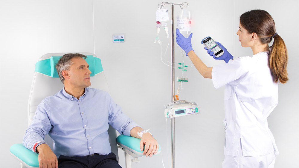 A imagem mostra um paciente sentado em uma cadeira, recebendo uma infusão de um dispositivo médico da B. Braun, enquanto uma enfermeira é vista configurando a dosagem de infusão no dispositivo.
