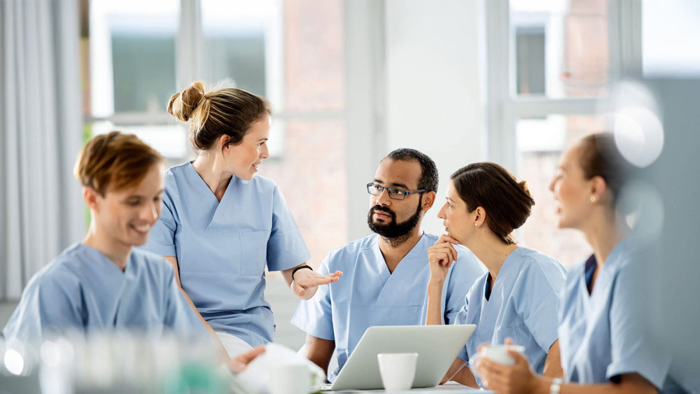 Equipe de saúde vestindo aventais azul-claro, sentada ao redor de uma mesa em um ambiente hospitalar bem iluminado, discutindo o atendimento ao paciente, com um laptop e xícaras de café sobre a mesa