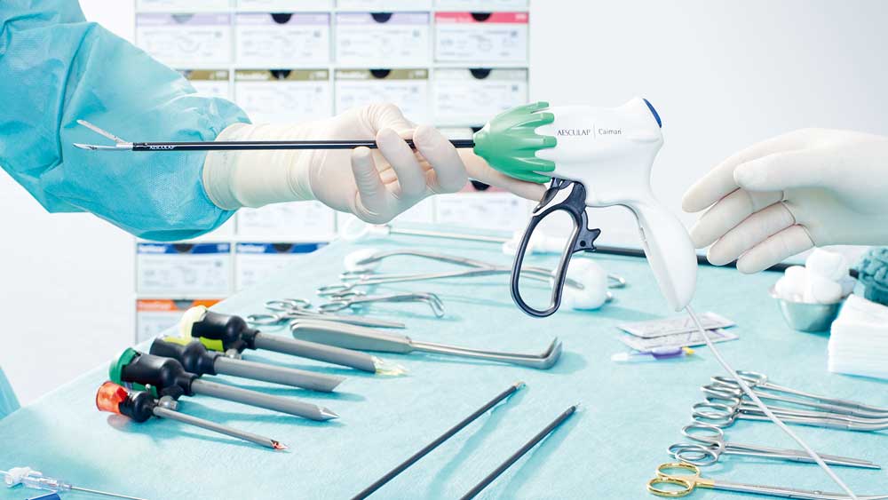 Um detalhe de uma situação cirúrgica minimamente invasiva: uma pessoa vestida com roupa cirúrgica entrega um Caiman a outra; ao fundo, há uma mesa com instrumentos cirúrgicos cuidadosamente dispostos para uma cirurgia