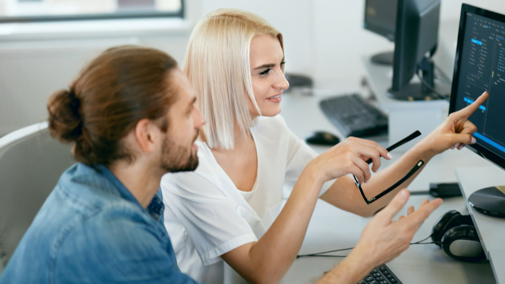 Dois profissionais de saúde estão sentados em frente a um computador. Ela mostra algo para ele na tela.
