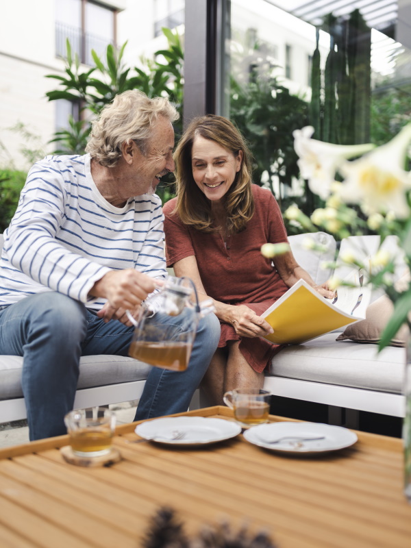 Um homem com um bule de chá e uma mulher com uma revista sentam-se em um sofá e riem