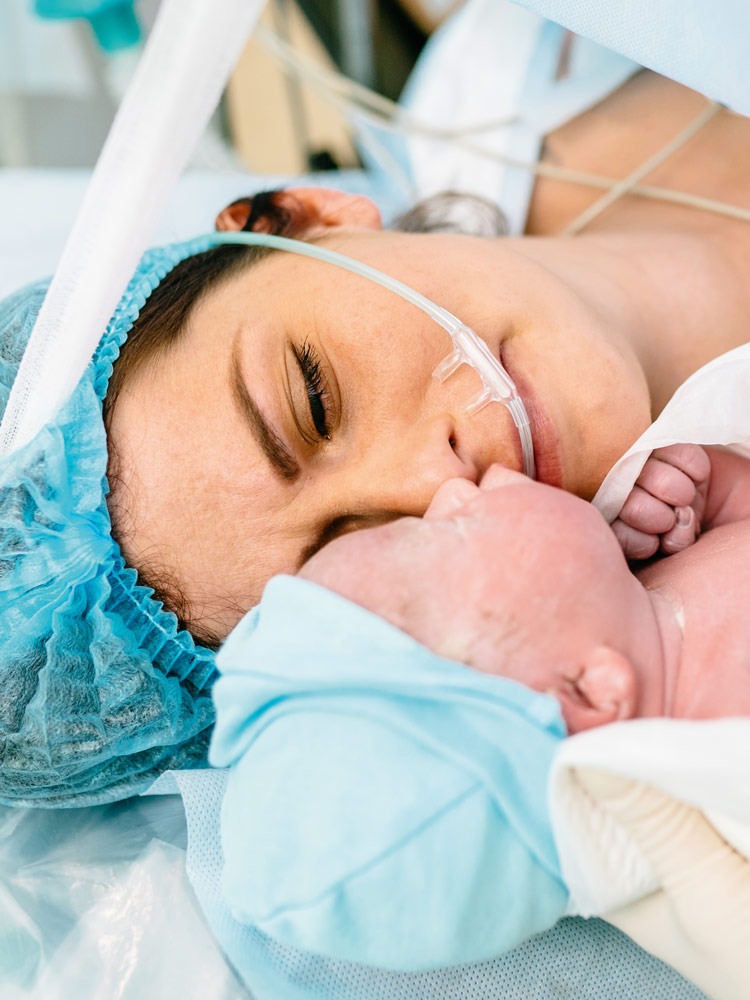 Uma mãe está deitada na sala de cirurgia após uma cesariana e olha para seu bebê recém-nascido, que uma enfermeira lhe mostra. Ela parece exausta, mas também aliviada. Ela está usando uma touca cirúrgica sobre os cabelos escuros, o recém-nascido está envolto em um pano branco e usa um gorro azul claro.