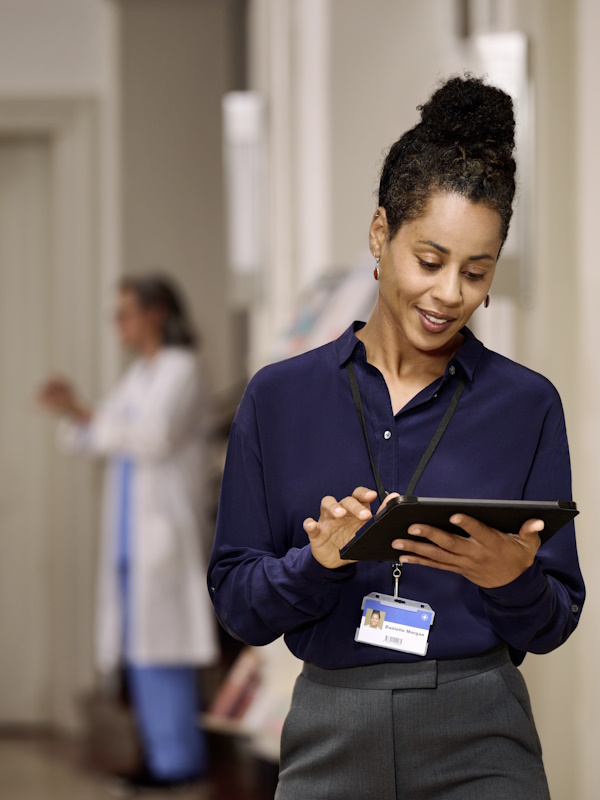 Uma mulher em primeiro plano, vestida com uma camisa azul escura e usando um crachá de identificação em um cordão, está parada em um corredor, segurando e olhando para um tablet. Ao fundo, é possível ver outra pessoa vestida com roupa médica.
