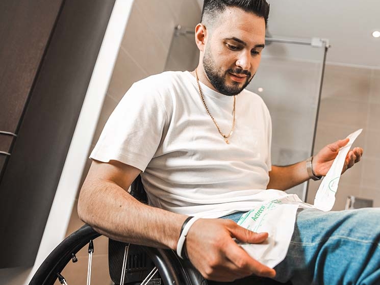 Um homem em uma cadeira de rodas, um banheiro ao fundo, segurando um cateter na mão