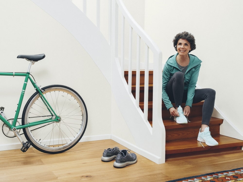 À direita, uma mulher está sentada nos degraus amarrando os sapatos; à esquerda, há uma bicicleta e outro par de sapatos