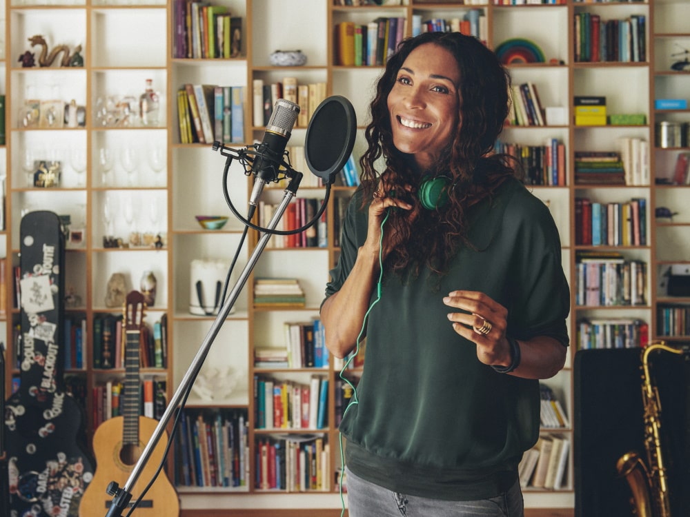 Em primeiro plano, uma mulher está diante de um microfone e sorri; ao fundo, há uma estante de livros cobrindo toda a parede