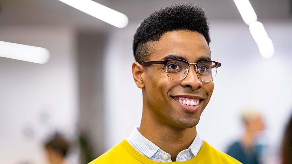 Close de um indivíduo sorridente com um top amarelo e óculos em um ambiente iluminado. 