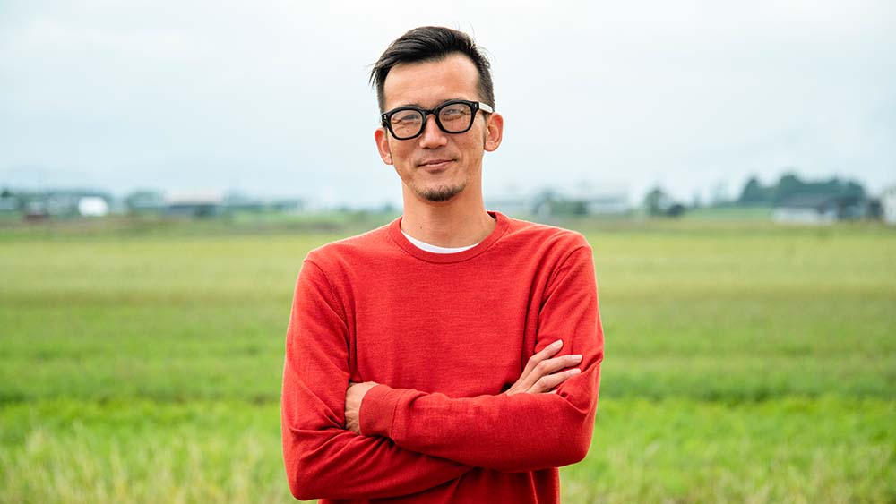 Uma pessoa usando óculos está de pé, confiante e com os braços cruzados em um vasto campo. 